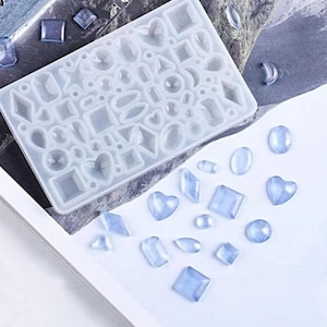 Pode incluir: Um molde de silicone translúcido retangular com várias cavidades geométricas, incluindo corações, círculos, quadrados e diamantes, acompanhado por várias peças de resina acabadas em azul claro com formas correspondentes.