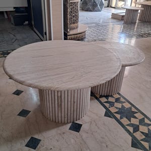 May include: A set of two round coffee tables made of beige travertine stone. Each table features a smooth, circular tabletop surface supported by a cylindrical base constructed from vertical, fluted stone rods arranged in a continuous pattern.