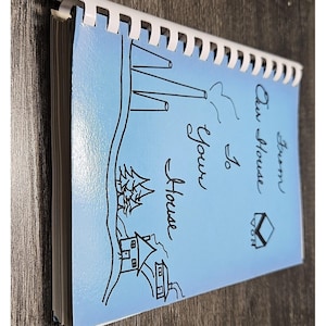May include: A light blue spiral-bound notebook with a black line drawing of a house, trees, and a chimney on the cover. The text "From Our House To Your House" is printed in black script, along with a small house-shaped logo containing the letters VON.