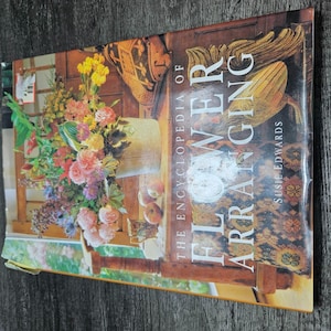 May include: A hardcover book titled The Encyclopedia of Flower Arranging by Susie Edwards. The cover features a colorful floral arrangement in a ceramic container, set on a wooden table with a patterned textile and two apples nearby.