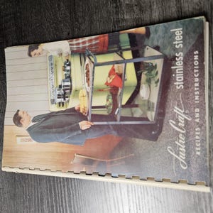 May include: A vintage spiral-bound cookbook titled Lustre Craft stainless steel recipes and instructions. The cover features a colour illustration of a person in a dark suit standing next to a yellow kitchen cart with food and cookware.