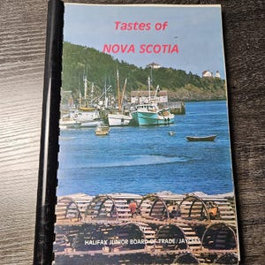May include: A spiral-bound cookbook titled Tastes of NOVA SCOTIA by HALIFAX JUNIOR BOARD OF TRADE/JAYCEES. The cover features a photograph of fishing boats docked in a harbour with a green hillside and buildings in the background and lobster traps.
