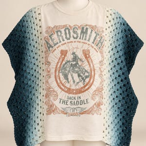 May include: A cream-colored t-shirt featuring a graphic of a cowboy on a horse inside a horseshoe with the text AEROSMITH, RIDIN' INTO TOWN ALONE BY THE LIGHT OF THE MOON, and BACK IN THE SADDLE. The sides have blue ombre crochet panels.