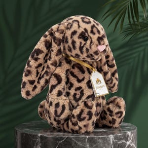 Pode incluir: Um peluche em forma de coelho com orelhas compridas, coberto com um padrão de leopardo em tons de bege e castanho escuro. O coelho tem um pequeno nariz cor-de-rosa, olhos pretos e uma fita dourada ao pescoço com uma etiqueta branca Jellycat.