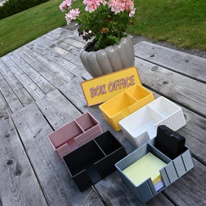 May include: A collection of five rectangular plastic desk organizers in yellow, white, pink, black, and gray, each featuring two compartments. A yellow sign with the text BOX OFFICE in pink letters is positioned behind the organizers.