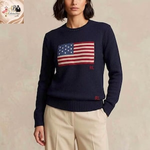 Può includere: Un maglione a maniche lunghe blu navy con girocollo caratterizzato da una grande bandiera americana lavorata a maglia sul petto. Il maglione presenta le lettere RL ricamate in rosso sotto la bandiera e vicino all'orlo inferiore, con pantaloni beige.