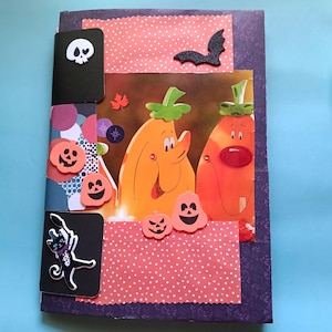 Puede incluir: Un cuaderno rectangular con temática de Halloween que presenta una cubierta de color púrpura oscuro con un patrón floral. La parte frontal muestra una ilustración central de dos calabazas naranjas, acentuadas con tiras de papel rosa con lunares, un murciélago negro, una calavera y un gato negro.