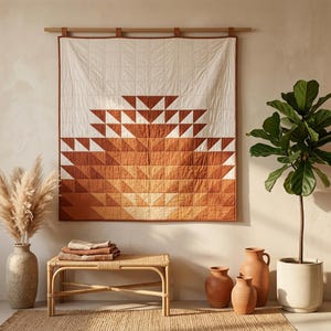 Op de afbeelding: Een vierkante wandquilt met een geometrisch patroon van driehoeken in tinten crème, roestoranje en terracotta. De quilt hangt aan een houten roede met stoffen lussen en heeft een doorgestikte rasterstructuur over het gehele oppervlak.