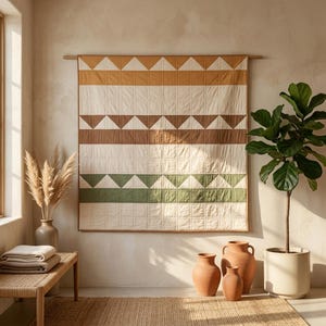 Op de afbeelding: Een rechthoekige wandquilt met horizontale banen van crèmekleurige, mosterdgele, bruine en saliegroene stof. Het ontwerp bevat herhalende driehoekspatronen en een rasterachtige doorgestikte textuur, opgehangen aan een houten roede.