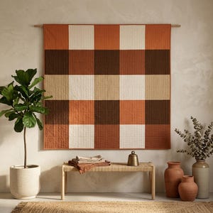 May include: A square wall hanging quilt featuring a large-scale buffalo check pattern in alternating blocks of terracotta orange, dark brown, and cream. The fabric has a visible quilted texture and is suspended from a horizontal wooden rod.