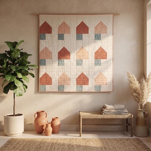 May include: A square wall hanging quilt featuring a repeating pattern of house shapes in shades of terracotta, peach, and muted blue on a cream background. The quilt is suspended from a wooden rod and displays a grid of twelve house motifs.