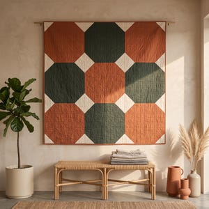 Puede incluir: Un edredón de pared cuadrado con un patrón geométrico de grandes octágonos en verde oscuro y naranja quemado, separados por pequeños cuadrados blancos. El edredón cuelga de una barra horizontal y muestra un patrón de costuras rectas.