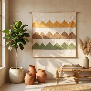 Puede incluir: Un edredón cuadrado para colgar en la pared con bandas horizontales de triángulos de color amarillo mostaza, marrón claro y verde salvia sobre un fondo crema, suspendido de una barra de madera. El edredón tiene costuras en cuadrícula y un borde fino marrón.