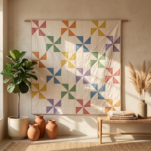 Könnte beinhalten: Ein quadratischer Wandquilt mit einem sich wiederholenden Windradmuster in den Farben Rosa, Orange, Gelb, Grün, Blau und Violett auf cremefarbenem Grund. Der Quilt ist an einer Holzstange befestigt und weist sichtbare Steppnähte auf.