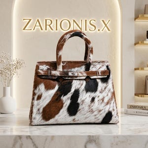 Puede incluir: Un bolso con estampado de piel de vaca con parches de pelo marrón, negro y blanco. El bolso tiene dos asas superiores, una correa horizontal en la parte delantera y un cierre de metal plateado. El texto ZARIONIS.X es visible al fondo.