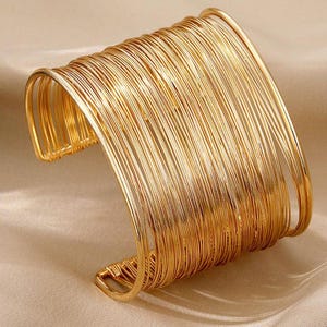 May include: A wide cuff bracelet constructed from numerous thin, polished gold-tone wire strands arranged in a parallel, multi-layered design. The open-ended circular structure features a continuous wrap of metallic wire, creating a textured, layered appearance with a reflective finish.