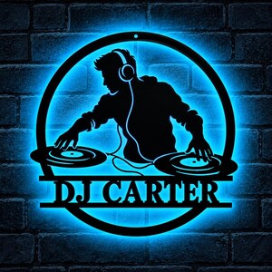 Könnte beinhalten: Ein kreisförmiges Wand-Schild aus schwarzem Metall mit der Silhouette eines DJs, der Kopfhörer trägt und zwei Plattenspieler bedient. Das Design wird durch hellblaues LED-Licht beleuchtet, und der Text DJ CARTER steht in fetten Buchstaben unten.