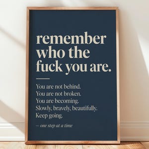 May include: A rectangular wall art print in a light wood frame featuring a dark navy blue background with cream-coloured text that reads: remember who the fuck you are. You are not behind. You are not broken. You are becoming. Slowly, bravely, beautifully. Keep going. — one step at a time.