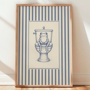 May include: A framed wall art print featuring a blue line drawing of a frog sitting on a toilet. The central illustration is set against a cream-colored background, which is framed by a border of vertical blue and cream stripes on the outer edge.