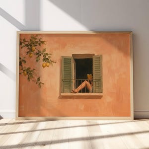 May include: A framed wall art print featuring a painting of a peach-colored stucco wall with a lemon tree branch on the left. A central open window with green shutters shows a person sitting on the sill reading a book with their legs extended.