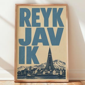 May include: A rectangular wall art print in a wooden frame featuring the word REYKJAVIK in large blue block letters. Below the text is a dark blue illustration of a city skyline with a tall church tower, mountains, and a boat on the water.