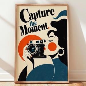May include: A framed poster featuring a stylized illustration of a person holding a black camera. The text Capture the Moment is printed in black at the top. The design uses a cream background with bold orange and dark blue accents throughout.