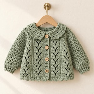 May include: A sage green long-sleeve cardigan featuring a scalloped collar and hem, vertical leaf-patterned crochet detailing, and four light brown wooden buttons down the center front. The garment is displayed on a wooden hanger.