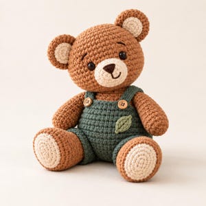Puede incluir: Un oso de peluche tejido a ganchillo en color marrón con el hocico y el interior de las orejas en color crema. El oso lleva un peto tejido a ganchillo en verde oscuro con dos botones de madera en los tirantes y una hoja verde claro en el pecho.