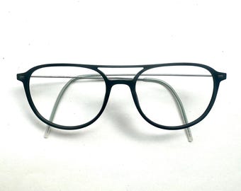 May include: A pair of matte black eyeglasses featuring a double bridge design. The frames have a rounded shape with thin, metallic silver-toned temples and clear, translucent temple tips. The overall construction is lightweight and minimalist.