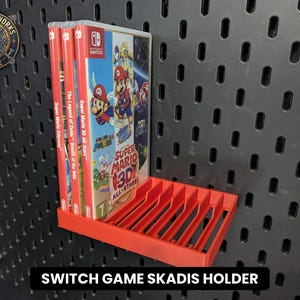 May include: A bright red plastic storage rack designed for a pegboard, featuring multiple vertical slots to hold Nintendo Switch game cases. Three game cases are currently inserted into the rack, which is mounted on a black pegboard wall panel.