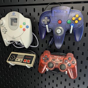 Peut inclure: Une collection de quatre manettes de jeu vidéo vintage montées sur un panneau perforé noir. L'ensemble comprend une manette Sega Dreamcast blanche, une manette Nintendo 64 violette translucide, une manette Nintendo NES rectangulaire et une manette Sony PlayStation rouge translucide.