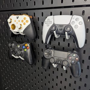 Puede incluir: Cuatro mandos de videojuegos montados en un panel perforado negro mediante ganchos de plástico blanco. La colección incluye un mando de Xbox blanco y dorado, un mando de Xbox 360 negro, un mando de PlayStation 5 blanco y un mando de PlayStation 4 negro.