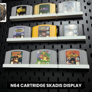 Puede incluir: Un expositor de pared de tablero perforado negro con tres filas de estantes de plástico blanco que sostienen nueve cartuchos de videojuegos Nintendo 64. Los cartuchos incluyen títulos como The Legend of Zelda, Donkey Kong 64 y Super Mario 64. Texto: N64 CARTRIDGE SKADIS DISPLAY.