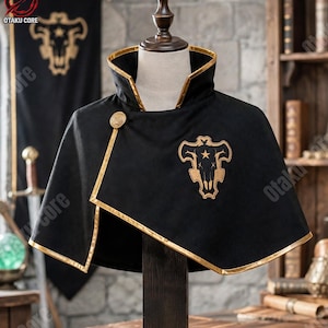 May include: A black cape with a high collar and gold trim, featuring a gold embroidered Black Bull emblem on the chest and a gold button closure. The cape is displayed on a mannequin torso, showcasing the structured fabric and detailed stitching.