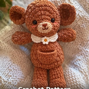 Puede incluir: Un animal de peluche tejido a ganchillo de color marrón con orejas redondas, ojos negros y un hocico color crema. Lleva un cuello tejido blanco con una flor de margarita central y tiene un pequeño bolsillo en la barriga. El texto Crochet Pattern es visible.