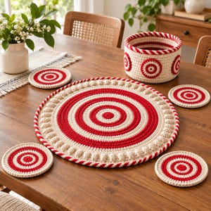 Puede incluir: Un conjunto de artículos de decoración para el hogar tejidos a ganchillo con un patrón circular en rojo y crema. La colección incluye un mantel individual redondo grande, cuatro posavasos redondos pequeños y una cesta cilíndrica con detalles circulares.