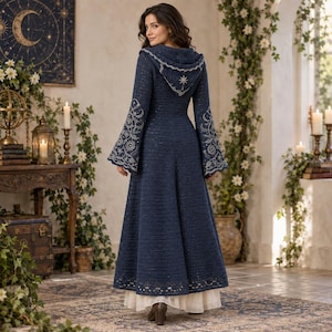 Könnte beinhalten: Ein bodenlanger, dunkelblauer Mantel mit Kapuze und langen, ausgestellten Ärmeln, die mit silbernen Stickereien von Himmelsmotiven wie Sternen und Monden verziert sind. Der Mantel hat eine gestrickte Textur und einen gewellten Saum über einem weißen Rock.