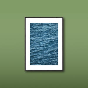 May include: A vertical rectangular wall art print featuring a high-angle view of deep blue ocean water with small, rippling waves. The print is encased in a thin black frame with a wide white mat border, presenting a clean and structured display.