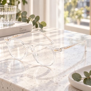 May include: Clear, round-framed glasses featuring a transparent acetate frame and thin, metallic gold-toned accents at the hinges. The eyewear has a classic circular silhouette with a bridge and temples that maintain a consistent, see-through aesthetic.