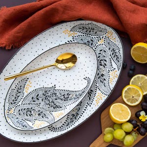 May include: Two white oval ceramic platters featuring a black and yellow paisley leaf pattern and black polka dots. A gold-toned metal spoon rests on the smaller platter. The dishes are arranged with sliced lemons and grapes nearby.