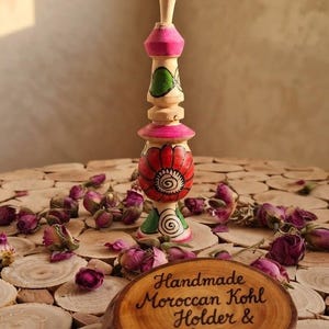 May include: A wooden kohl holder featuring a tall, slender shape with a red, pink, and green painted floral design. A small wooden plaque in front displays the text: Handmade Moroccan Kohl Holder & Boho Decor, written in black ink.