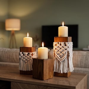 May include: Three square wooden candle holders of varying heights, each topped with a cream-colored pillar candle. Two of the holders are wrapped in white macrame cord with a knotted fringe design. The wood has a dark, natural grain finish.