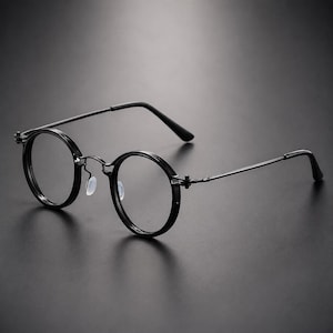 May include: A pair of black round-frame glasses featuring thin metal temples, a slim bridge, and clear lenses. The eyewear includes adjustable translucent nose pads and a minimalist construction with visible hinges connecting the frame to the arms.