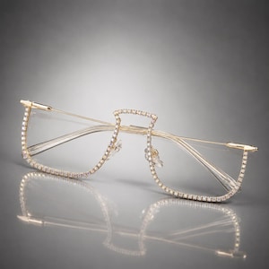 Puede incluir: Gafas sin montura con estructura de metal en tono dorado, adornadas con brillantes transparentes a lo largo de los bordes exteriores y el puente. El diseño incluye varillas finas doradas y plaquetas nasales transparentes.
