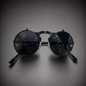 Puede incluir: Gafas de sol redondas con lentes circulares negras y una montura negra con un puente distintivo. El diseño incluye un mecanismo abatible en la parte superior de cada lente, lo que permite levantar las lentes oscuras de forma independiente.