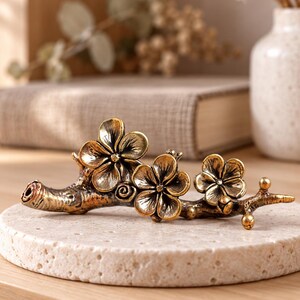 May include: A metallic, antique-gold coloured brush rest shaped like a tree branch with three blooming flowers. The object features a textured, weathered finish with dark highlights in the crevices, showcasing a detailed floral design.