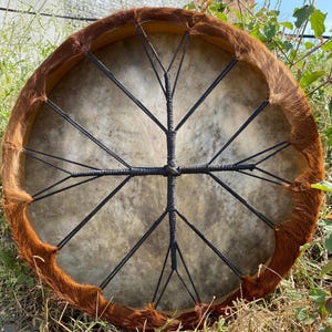 Op de afbeelding: Een ronde frametrommel met een lichtgekleurd, gevlekt dierenhuid trommelvel. De rand is omwikkeld met bruine, getextureerde vacht. Een zwart koord is in een kruispatroon over de achterkant van het vel gespannen om het aan het frame te bevestigen.