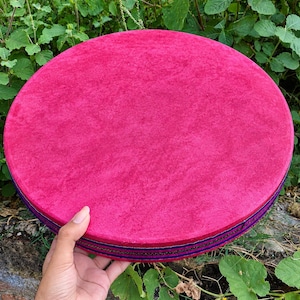 May include: A circular, flat object with a vibrant magenta, velvet-like top surface. The side edge features a decorative trim with horizontal stripes in dark blue, black, and gold, creating a distinct border around the round, pink frame.