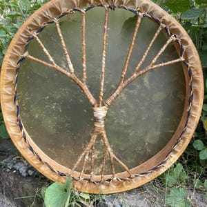 May include: A circular frame drum featuring a translucent, light brown drumhead stretched over a wooden hoop. The drumhead is secured with a decorative lacing pattern that forms a tree-like structure, using thin, light-coloured cordage.