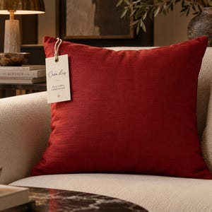 May include: A square, deep red throw pillow with a visible woven texture. A rectangular off-white paper tag is attached to the top corner with a light-colored string. The tag features the text Casa Luz, Silk Linen, and Handcrafted.